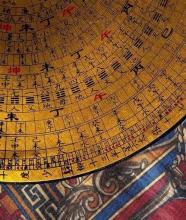  A close-up of a traditional Chinese Feng Shui Luo Pan compass, featuring golden concentric rings inscribed with black and red Chinese characters and symbols, resting on a colorful patterned fabric.          Ask ChatGPT