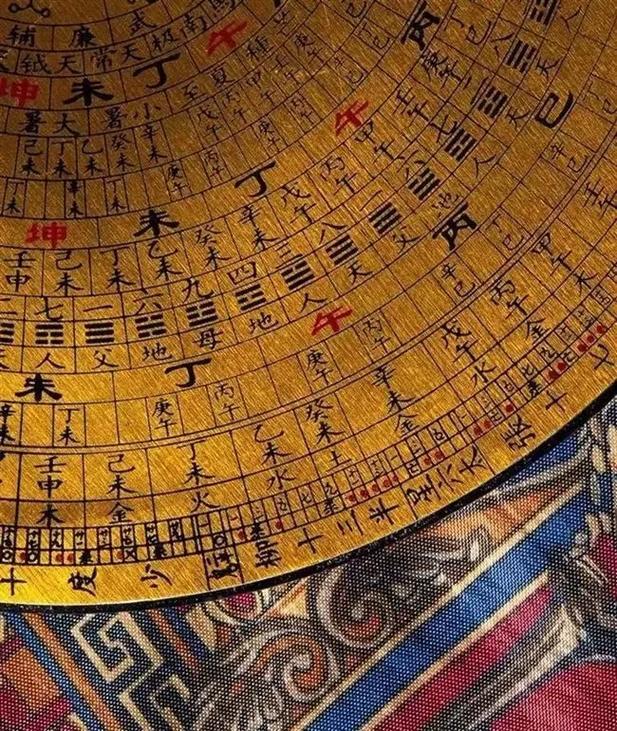  A close-up of a traditional Chinese Feng Shui Luo Pan compass, featuring golden concentric rings inscribed with black and red Chinese characters and symbols, resting on a colorful patterned fabric.          Ask ChatGPT