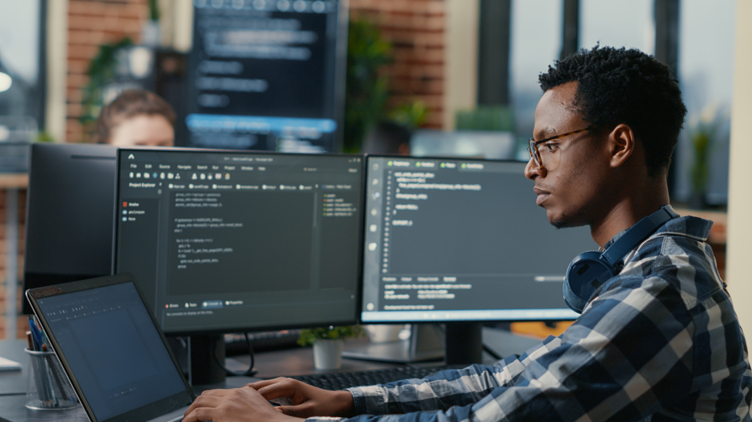 an employee siting in his cubicle, using ai to vibe code software development on one screen on other another ai program running to check the given code for errors.