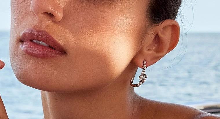 A woman by the ocean, showcasing diamond earrings, with the sea in the background.